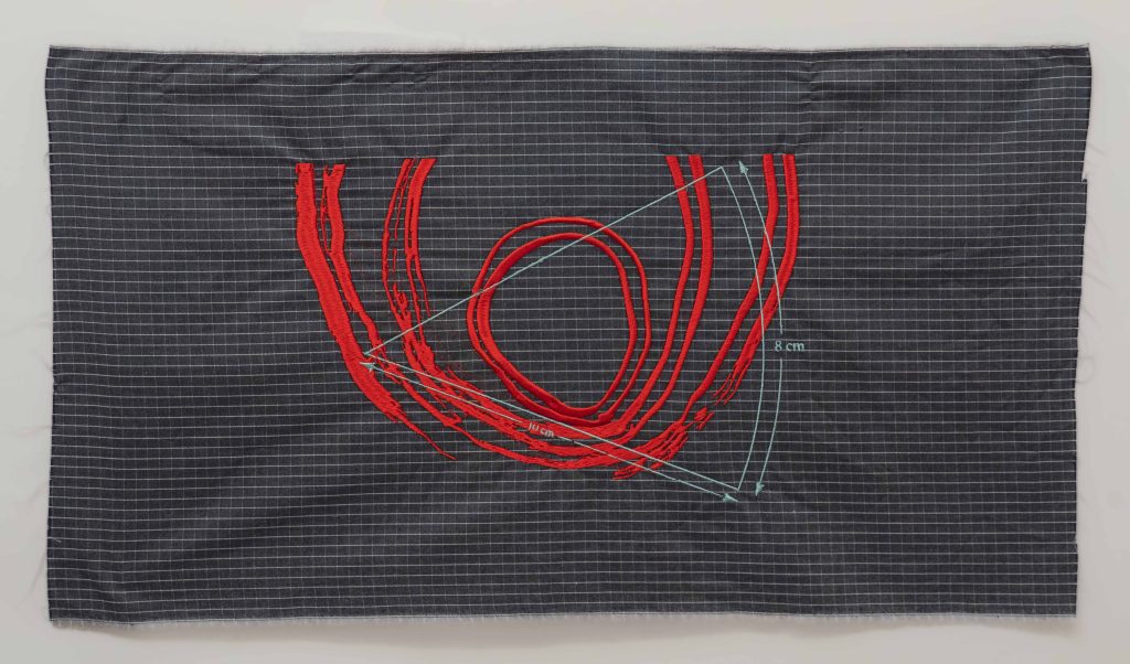 The image shows a photograph of machine embroidery on a densely checked unhemmed polycotton fabric with a dark grey background and white lines. The embroidery is made up of thick bright red, ever increasing, yet disintegrating circles and half circles that resemble the inner world of an oyster or fingerprint stitched in silky satin thread. This contrasts with a geometric triangular shape that resembles a line drawing of a slice of pie or cheese, with measurements of 10cm and 8cm stitched over the top of the red circles in an eggshell blue thread.