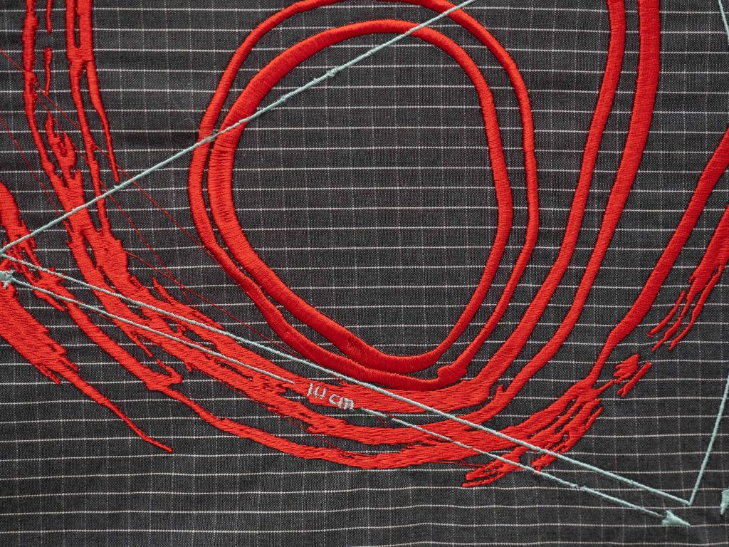 The image shows a photograph of machine embroidery on a densely checked polycotton fabric with a dark grey background and white lines. The embroidery is made up of thick bright red, ever increasing, yet disintegrating circles and half circles that resemble the inner world of an oyster or fingerprint stitched in silky satin thread. This contrasts with a geometric triangular shape with a measurement of 10cm stitched over the red circles in an eggshell blue thread.