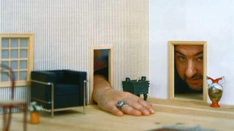 A hand reaches through a miniature doorway into a small-scale diorama of a living room, resting on the floor and wearing a blue ring on the third finger. To one side sits a blue armchair, with a wicker dining chair blurred in the foreground. Through a second doorway, a man’s face peers into the model. There is a red and white vase to the right of the doorway, oddly large in relation to the rest of the room.