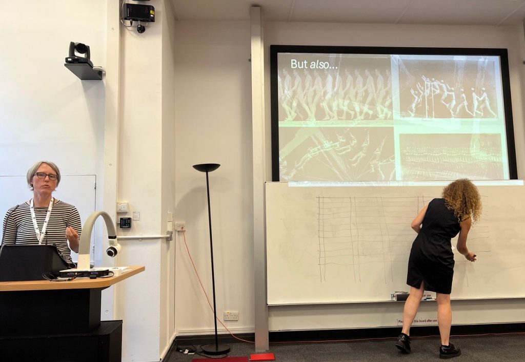 The photograph shows a view of the front of a typical lecture theatre or classroom. On the right a projection screen displays 19th-century motion-study photography and chronophotography illustrating human locomotion phases of upright male figures, notably featuring works by Eadweard Muybridge and Étienne-Jules Marey. These were the foundation of 'time and motion' studies by which awkward or redundant movements could be eliminated, tool layouts redesigns, fatigue reduced, and factory assembly-line efficiency maximised. 
Beneath the projected image is a whiteboard. A pale skinned woman in a black sleeveless knee height dress with red curly hair is drawing a chart on the whiteboard. She is leaning, a little awkwardly, to the right in order to bend to reach the extent of the board. On the left is a desk with a visualiser and lectern. A pale skinned grey haired woman with black rimmed glasses and a lanyard is speaking and making hand gestures to an invisible audience in front of the desk.