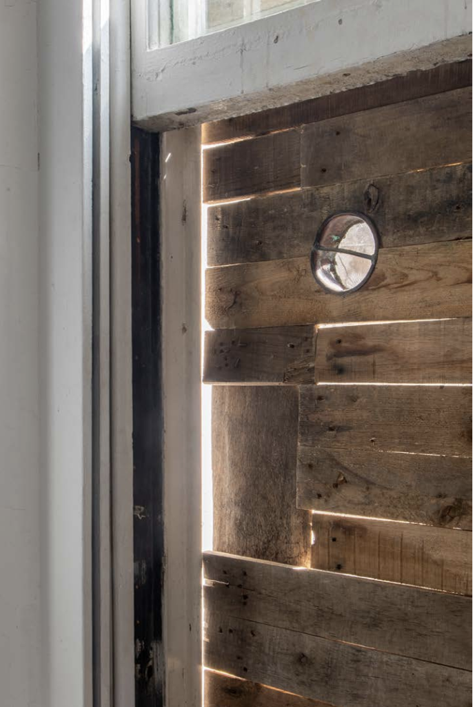 the exterior of a white sash window is boarded up with rough wooden planks, thin strips of daylight are visible through the gaps in the boards, and in the midst of them is a small round glass panel, with a metal rim along the diameter and across the centre.
