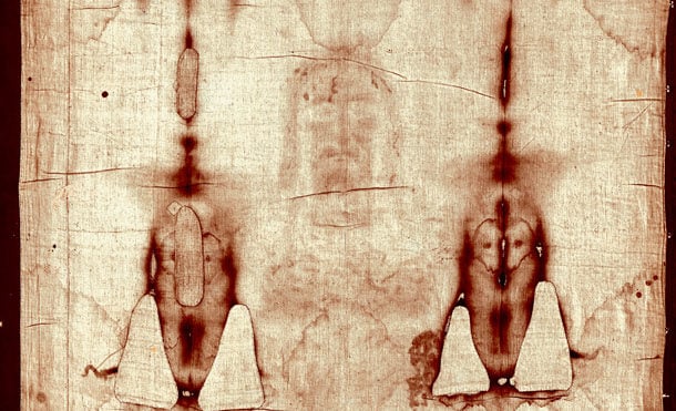 Sepia-toned photograph of the Shroud of Turin, showing front and back impressions of a human figure, faintly visible across the length of cloth.